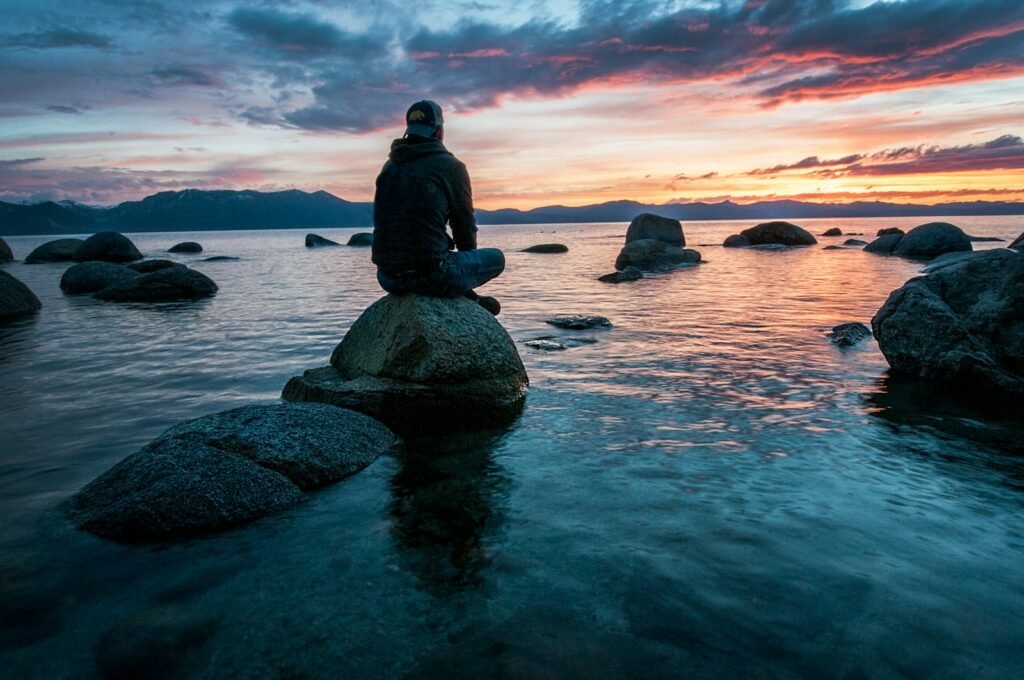 A man sitting on a rock surrounded by water, thinking about the benefits of meditation