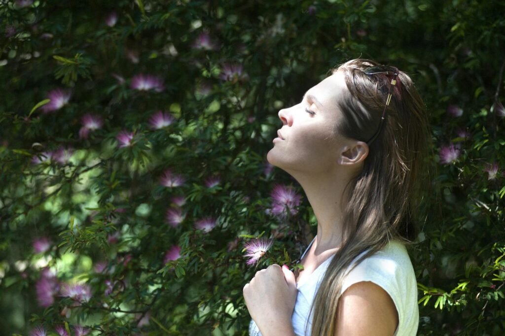 A woman closing her eyes against sunlight standing near a purple petaled flower plant for stress management