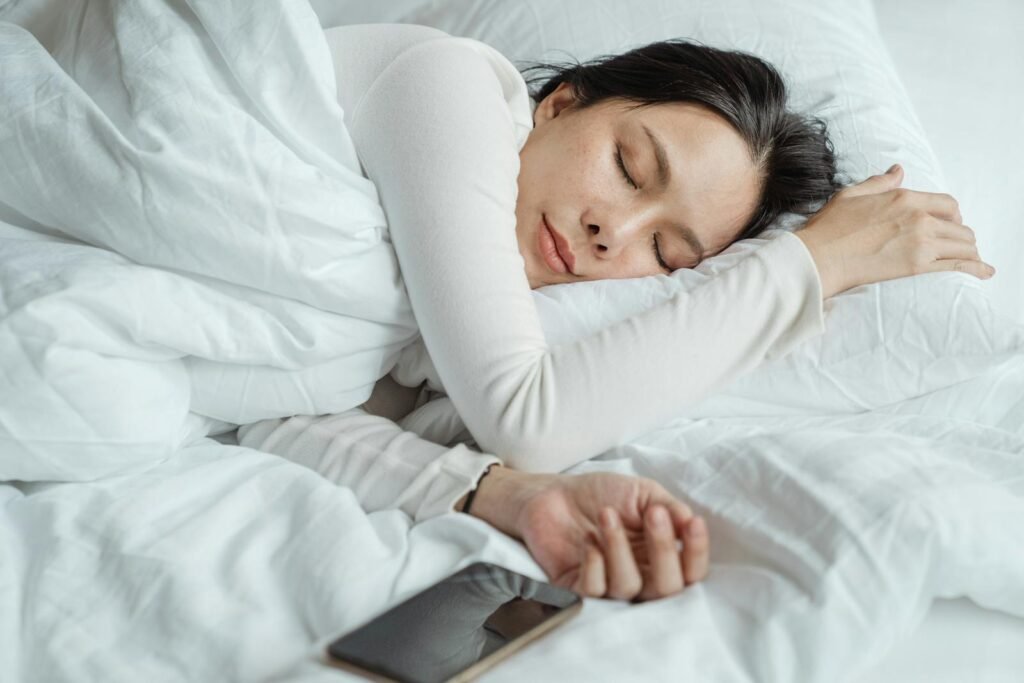 Calm female wearing white pajamas sleeping in a comfortable bed with white sheets near a modern mobile phone in the morning