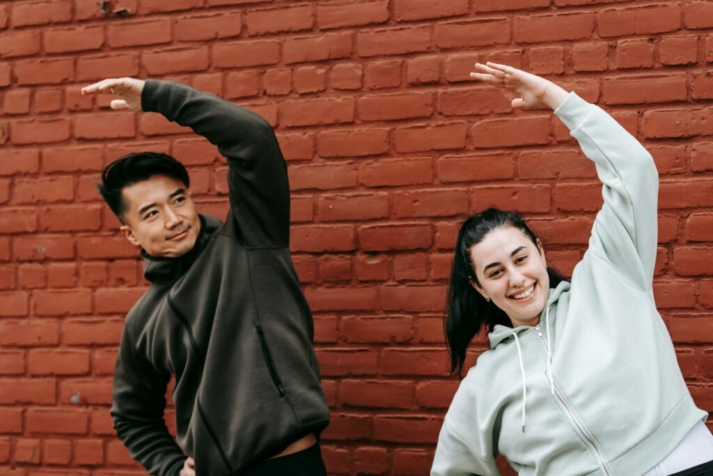 Positive young obese woman and Asian male trainer in sportswear warming up before outdoor training standing near brick wall