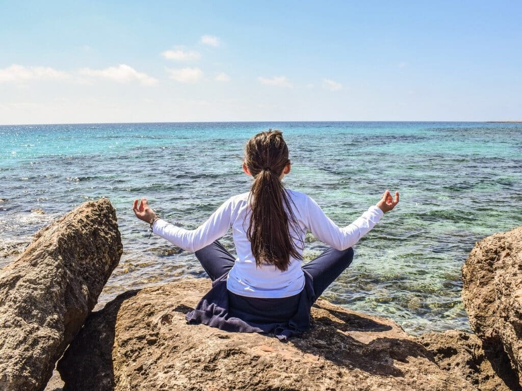 A girl sitting crossed-legged on rocks using meditation techniques to find inner peace by the sea