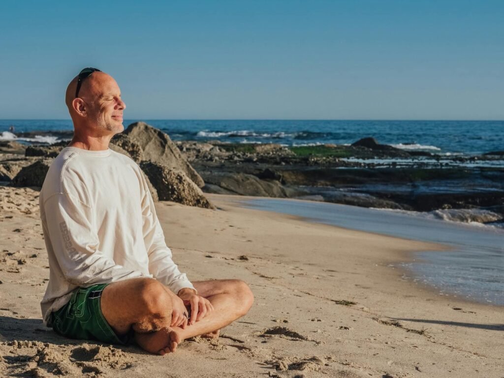 A bald man smiling while sitting in lotus pose on the beach