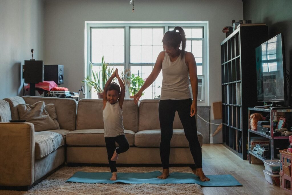 Entire body-positive mother teaching her child to perform Tree Pose on the mat in the cozy living room at home.